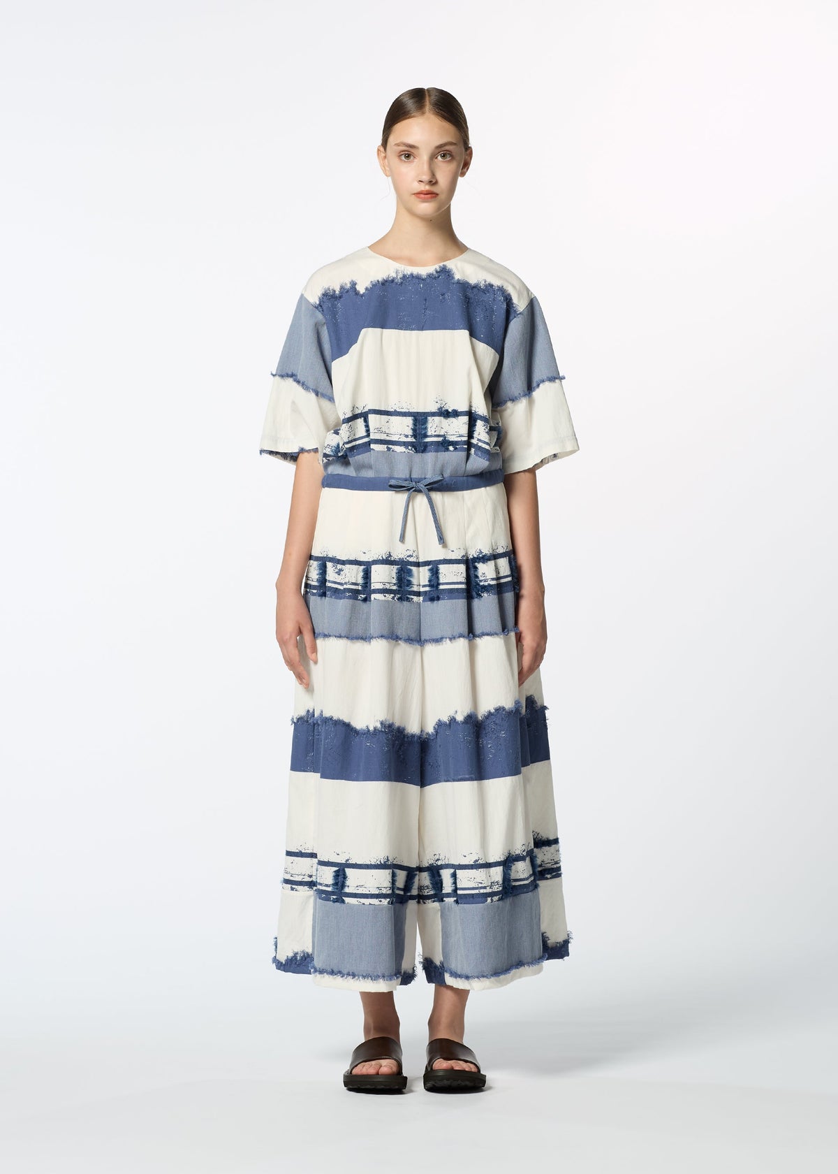 A person stands facing forward in the HaaT KASANEGAKE, a loose-fitting, short-sleeved, ankle-length dress with wide blue and white stripes and a layered design. They wear black sandals and have their hair pulled back against a white background. view 6 of 8