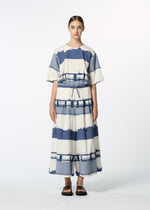 A person stands facing forward in the HaaT KASANEGAKE, a loose-fitting, short-sleeved, ankle-length dress with wide blue and white stripes and a layered design. They wear black sandals and have their hair pulled back against a white background.