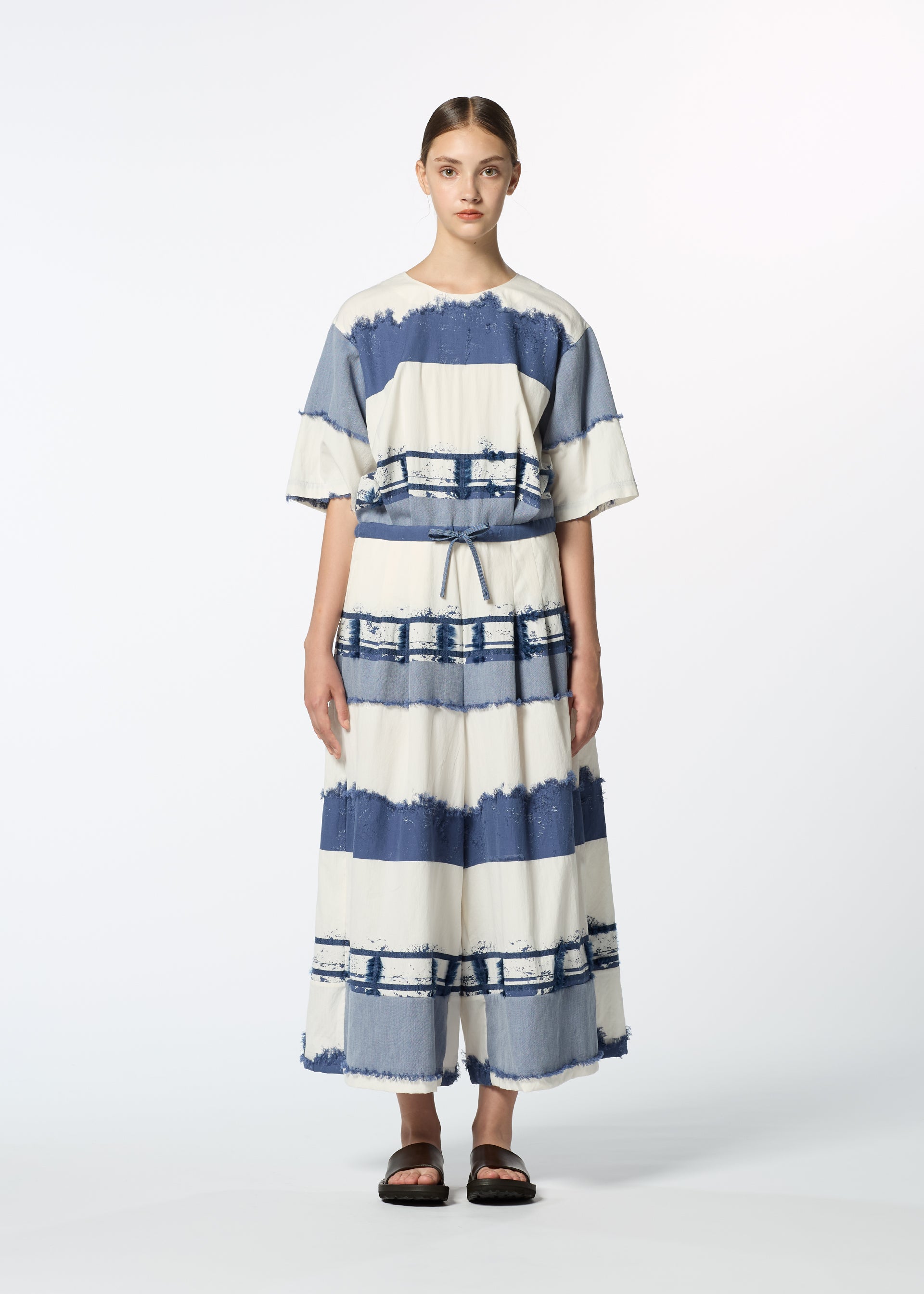 Zoomed view A person stands facing forward in the HaaT KASANEGAKE, a loose-fitting, short-sleeved, ankle-length dress with wide blue and white stripes and a layered design. They wear black sandals and have their hair pulled back against a white background.