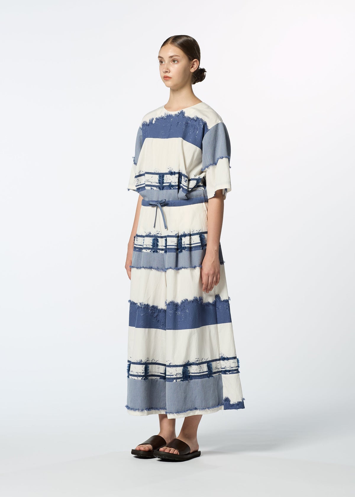 A woman stands against a plain background wearing HaaT's KASANEGAKE dress, featuring loose short sleeves, wide blue and white stripes, a matching cinched waist, and textured fabric. Her hair is tied back and she's in black sandals. view 7 of 8