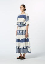 A woman stands against a plain background wearing HaaT's KASANEGAKE dress, featuring loose short sleeves, wide blue and white stripes, a matching cinched waist, and textured fabric. Her hair is tied back and she's in black sandals.
