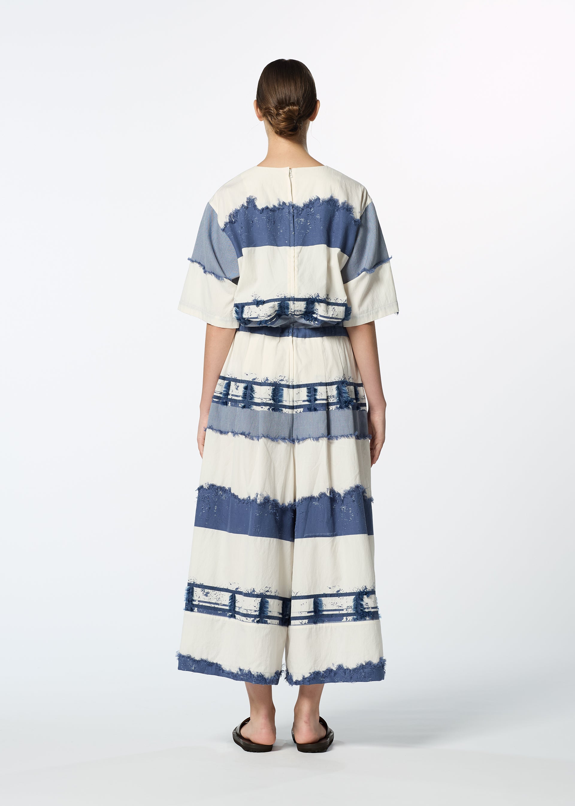 Zoomed view A person stands facing away, wearing HaaT's KASANEGAKE set—a loose, short-sleeved top and wide-legged pants in blue and white tie-dye stripes. The textured fabric and layered design add depth, paired with black sandals for a finished look.