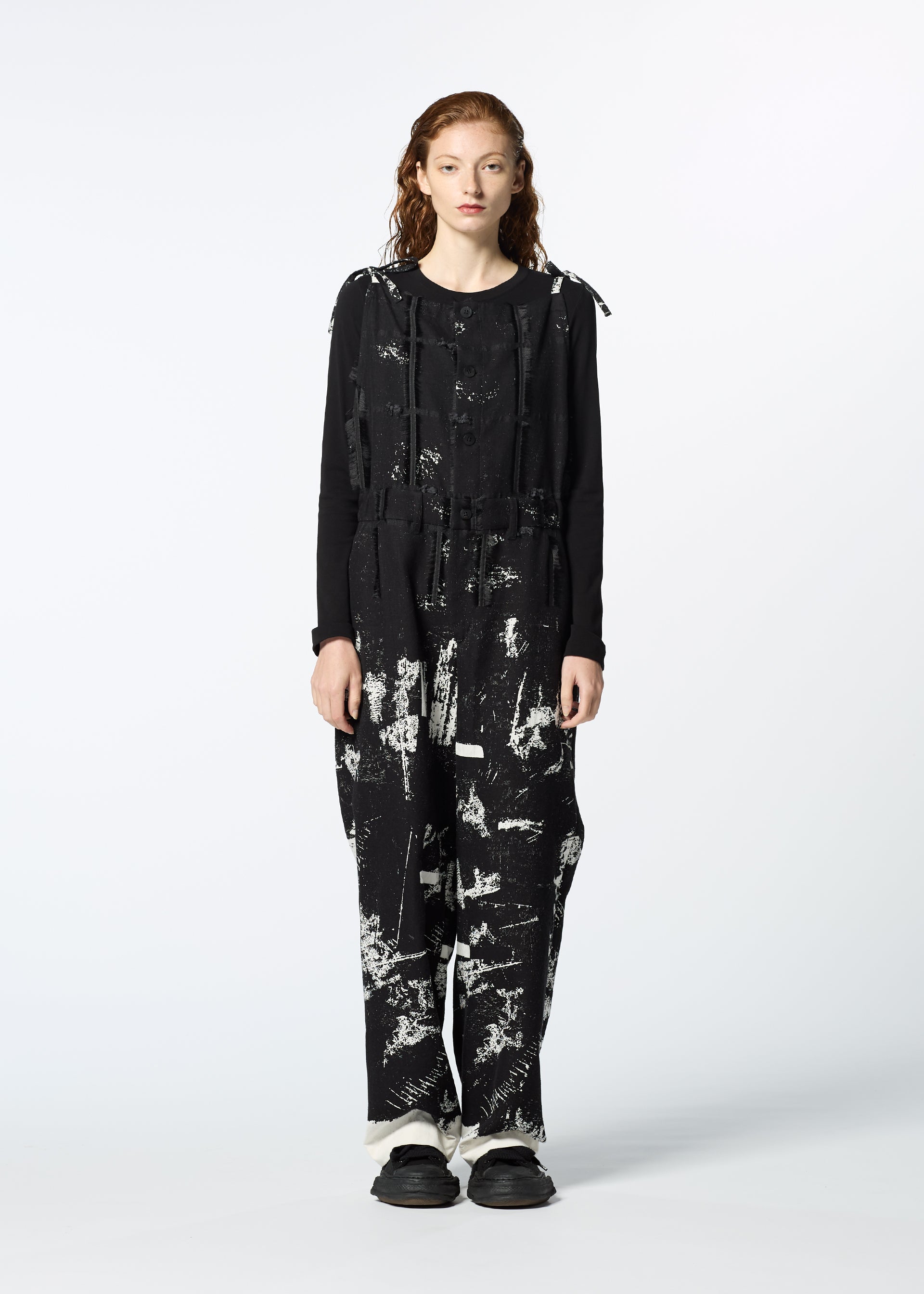 Zoomed view A person with long, wavy hair faces forward in the KOKUYU jumpsuit by HaaT, featuring expressive jacquard with white abstract paint splatters, loose-fitting pants, and black shoes against a plain white background.