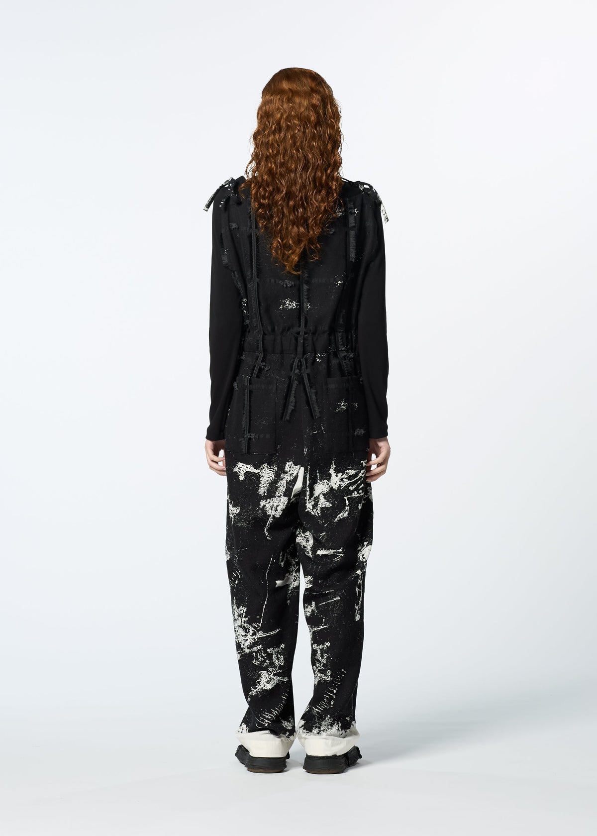 A person with long curly hair stands facing away, wearing HaaT’s KOKUYU jacquard jumpsuit over a black long-sleeve top and white-soled shoes, showcasing its expressive textile design against a plain light background. view 8 of 8