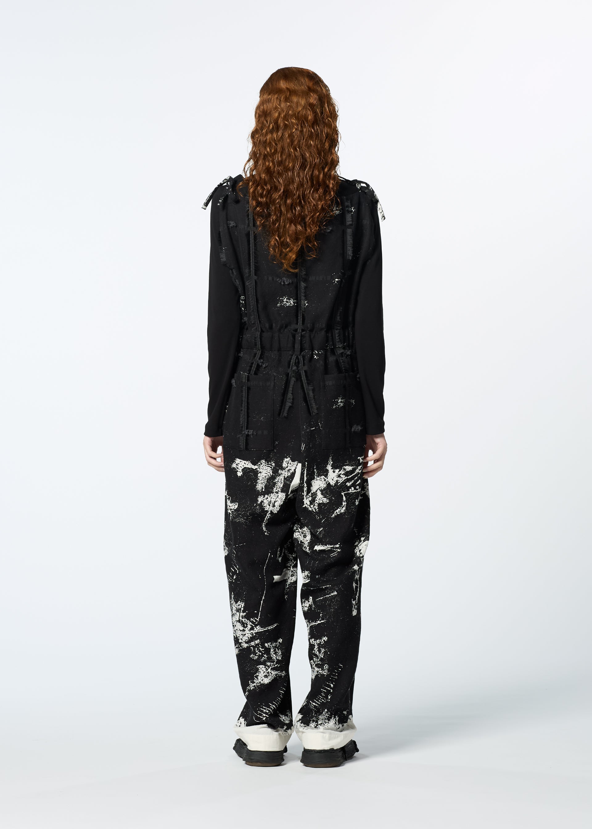 Zoomed view A person with long curly hair stands facing away, wearing HaaT’s KOKUYU jacquard jumpsuit over a black long-sleeve top and white-soled shoes, showcasing its expressive textile design against a plain light background.