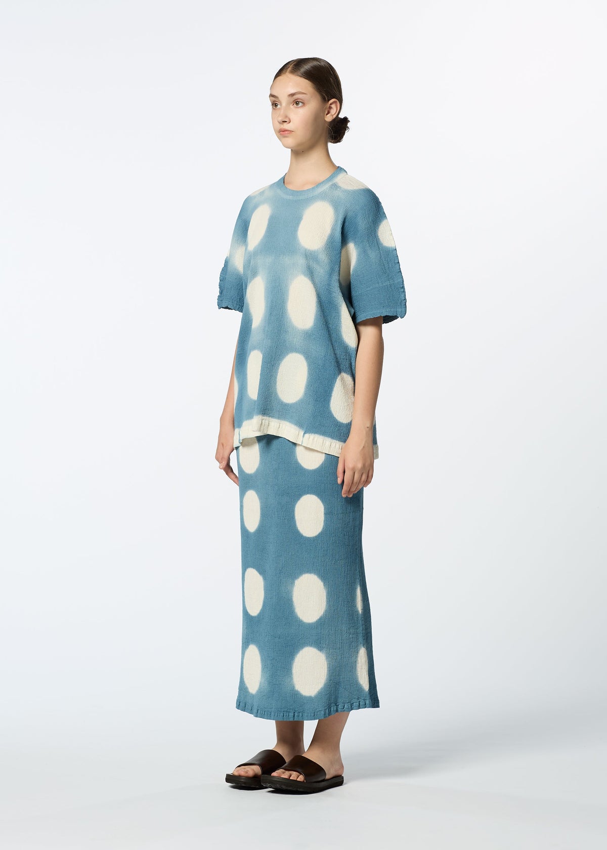 A woman in black sandals stands against a white background wearing HaaT’s KYO CHIJIMI ITAJIME: a loose-fitting blue short-sleeve top and matching long skirt with large white polka dots, crafted using the traditional Itajime technique. Her hair is pulled back. view 7 of 8