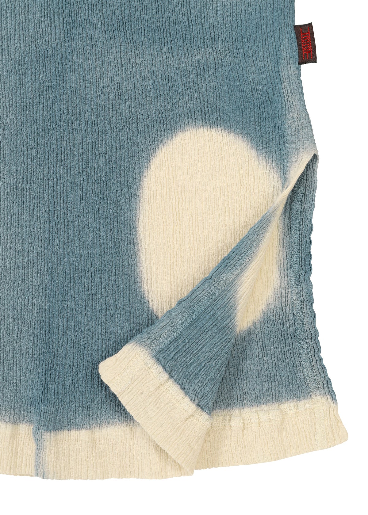 A close-up of the HaaT KYO CHIJIMI ITAJIME short-sleeve top highlights its blue and white textured fabric, crafted with the Itajime dyeing technique. A large white circle and a folded corner show the reverse, with a small red tag near the edge. view 5 of 8