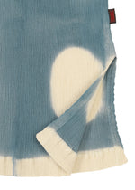 A close-up of the HaaT KYO CHIJIMI ITAJIME short-sleeve top highlights its blue and white textured fabric, crafted with the Itajime dyeing technique. A large white circle and a folded corner show the reverse, with a small red tag near the edge.