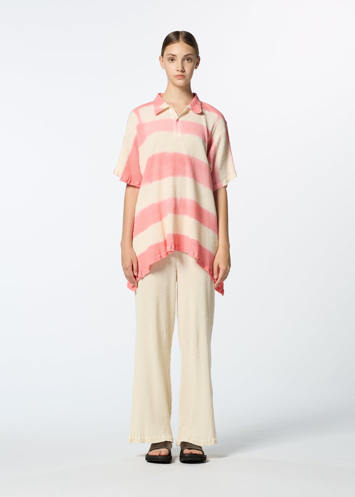 A person stands facing forward wearing HaaT's KYO CHIJIMI ITAJIME, a loose collared short-sleeve top with pink and cream stripes, paired with cream wide-leg pants and black sandals against a plain light background. view 6 of 8