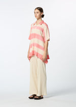 A woman stands against a plain background, wearing a loose HaaT KYO CHIJIMI ITAJIME collared short-sleeve top with pink and white stripes, wide cream-colored pants, and black sandals. Her hair is tied back as she faces slightly to the side.