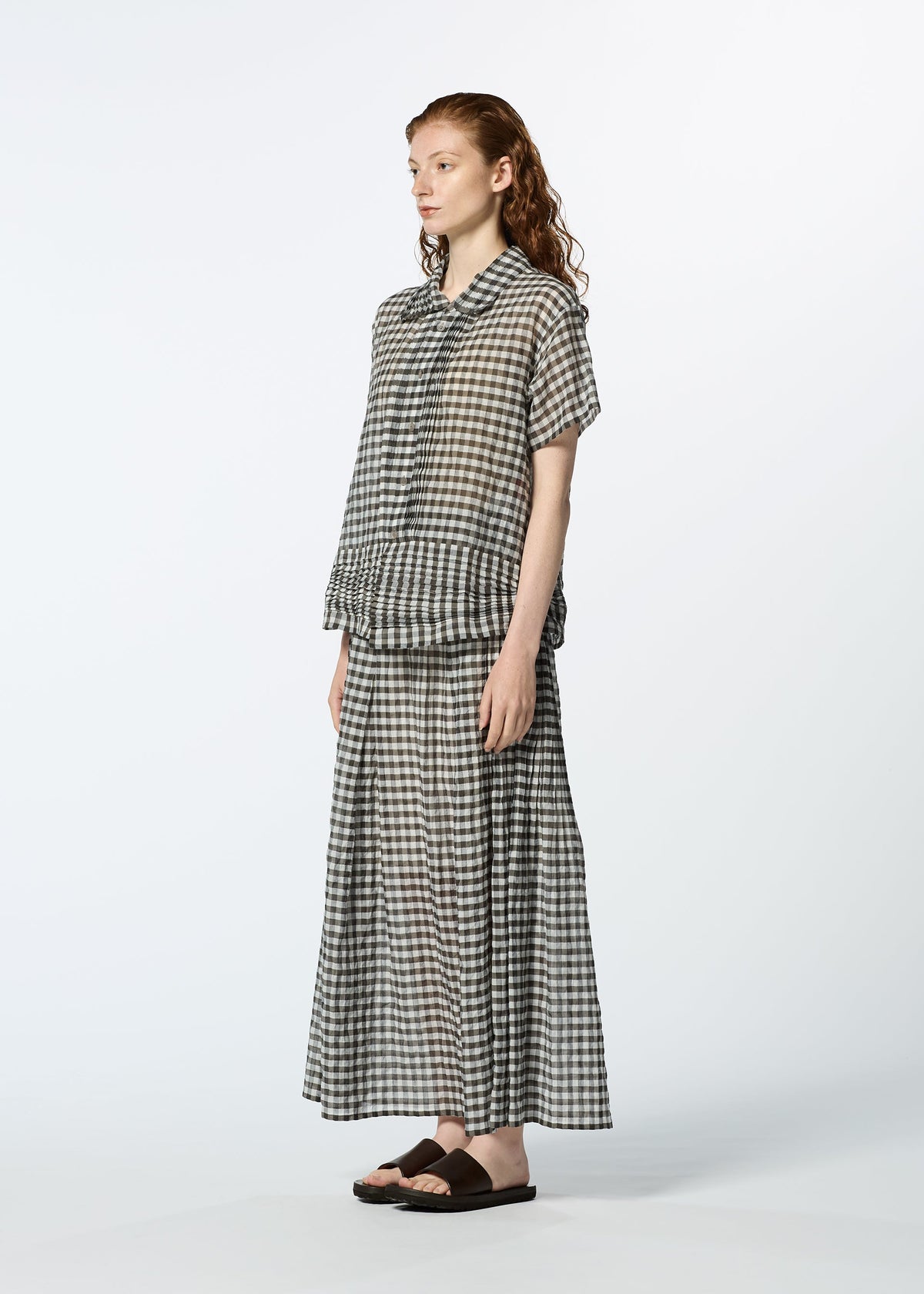 A person with long, wavy, reddish hair wears HaaT’s 2 SIDE PINTUCKING matching short-sleeve gingham shirt and ankle-length skirt with black sandals, standing against a plain white background. view 7 of 9