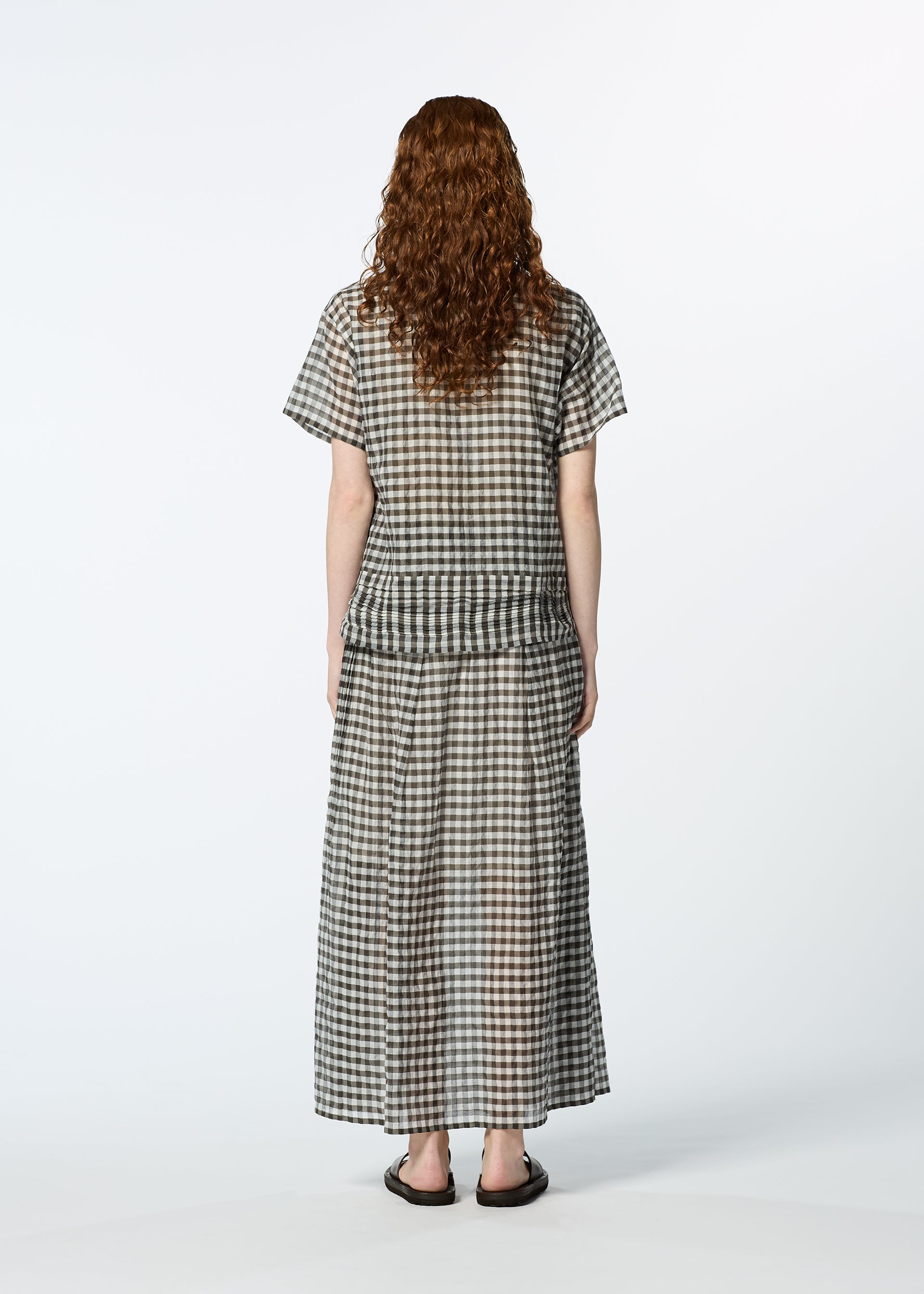 Zoomed view A person with curly, shoulder-length hair stands facing away, wearing HaaT's 2 SIDE PINTUCKING black and white gingham shirt and matching long skirt with dark sandals against a plain white background.