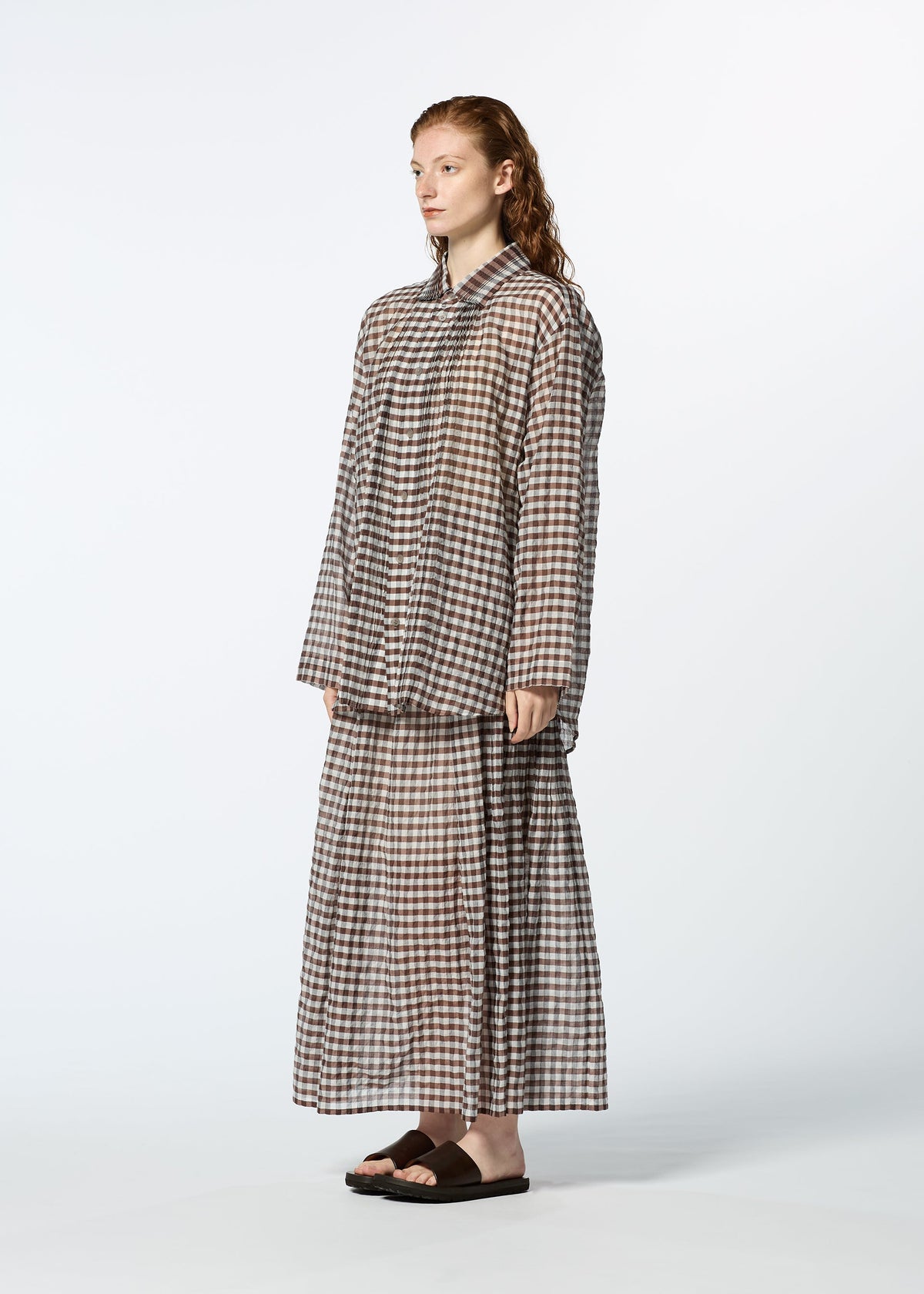 A person stands sideways wearing HaaT's 2 SIDE PINTUCKING brown and white gingham long-sleeve shirt and matching skirt with black slide sandals against a plain light background. view 7 of 8