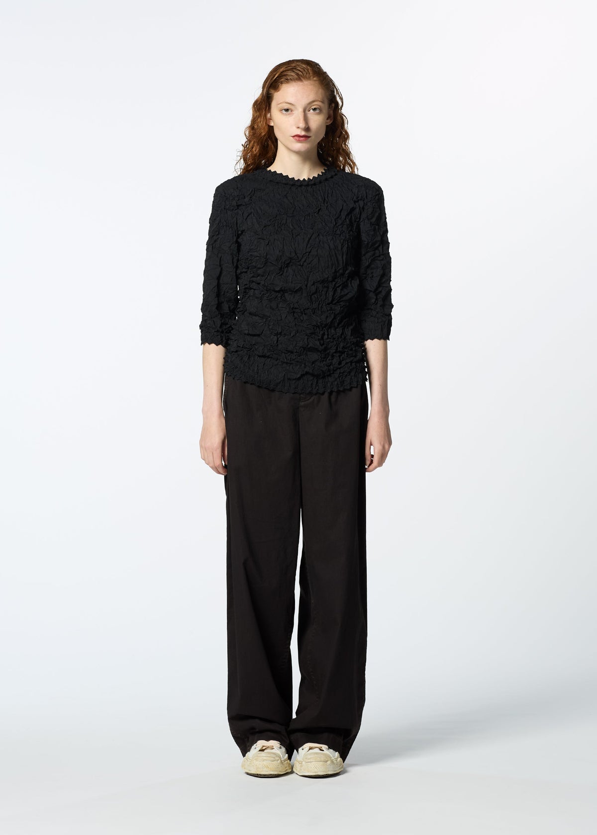 A person with long curly hair stands facing forward, wearing the textured black KUMO SHIBORI SHIRT by HaaT with three-quarter sleeves, loose black pants, and beige sneakers against a plain white background. view 7 of 9