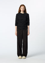 A person with long curly hair stands facing forward, wearing the textured black KUMO SHIBORI SHIRT by HaaT with three-quarter sleeves, loose black pants, and beige sneakers against a plain white background.