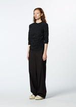 A person with long, wavy red hair stands in a studio wearing the KUMO SHIBORI SHIRT by HaaT, styled with loose black pants and white sneakers, facing slightly to the side against a plain white background.