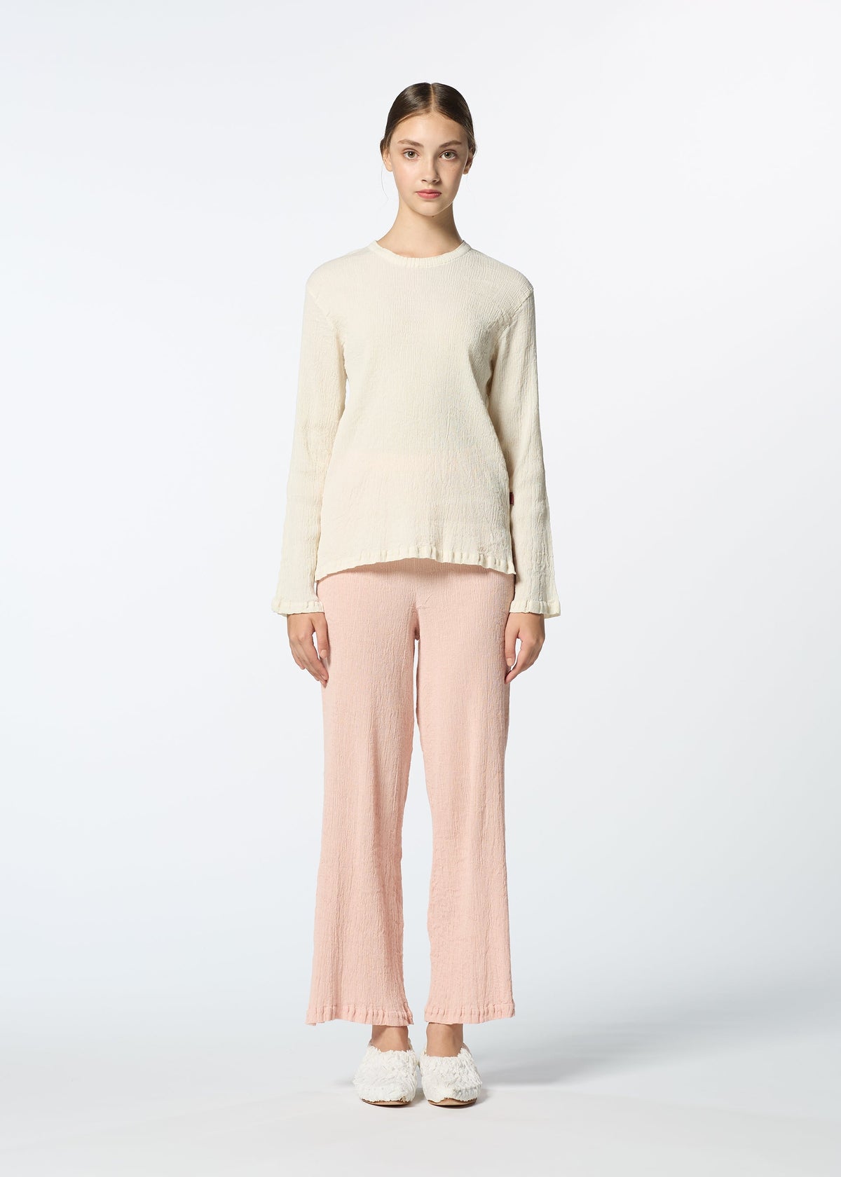 A young woman stands against a white background, wearing HaaT's cream KYO CHIJIMI FEBRUARY round-neck top, light pink wide-leg pants, and white slip-on shoes. Her hair is tied back as she looks straight at the camera. view 6 of 9