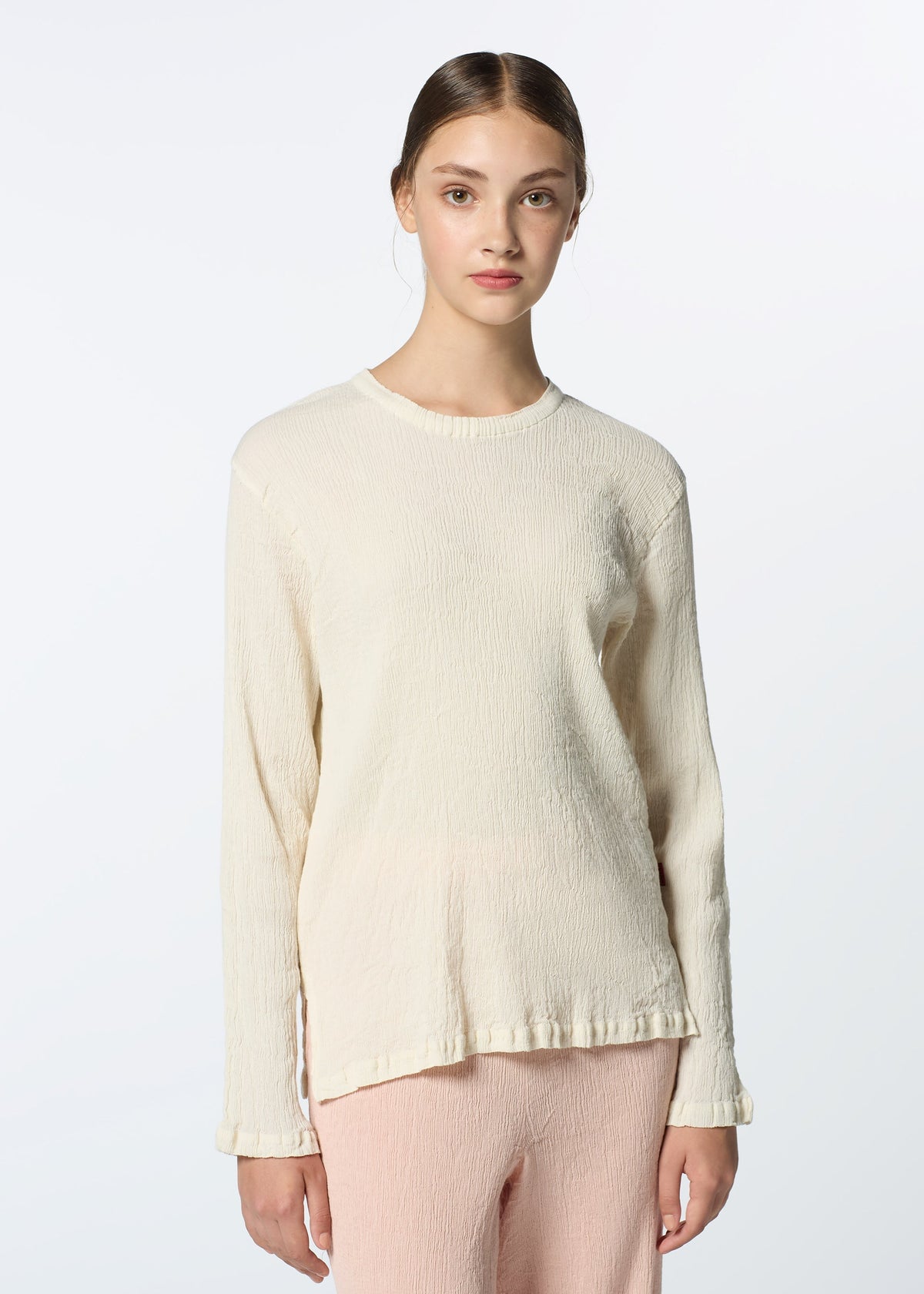 A young woman with straight brown hair parted in the middle wears HaaT’s KYO CHIJIMI FEBRUARY round-neck top and light pink pants, standing against a plain, light background, facing forward with a neutral expression. view 9 of 9