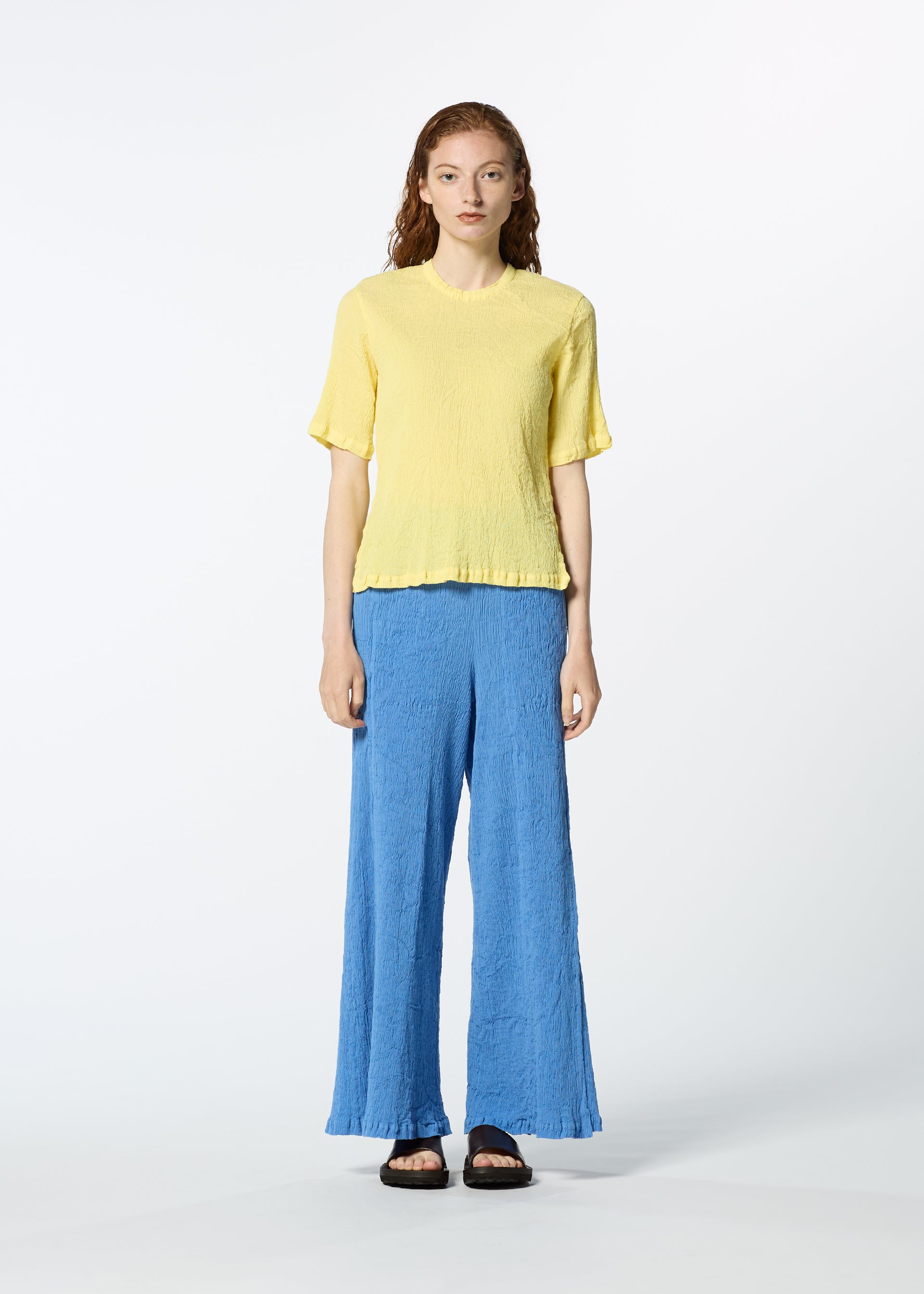 Zoomed view A person stands against a plain white background wearing a textured yellow KYO CHIJIMI MAY round-neck top from HaaT, loose blue wide-leg pants, and black sandals.