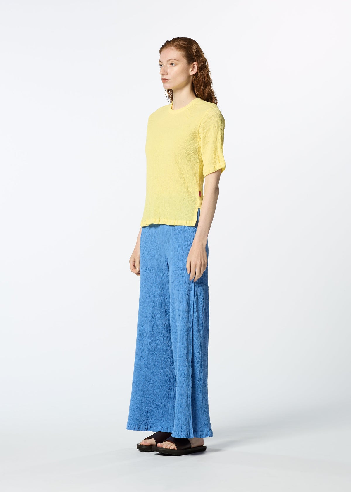 A person with long, wavy hair and a neutral expression stands against a white background, wearing HaaT's KYO CHIJIMI MAY textured yellow round-neck top in ultra-fine cotton, loose blue pants, and black sandals. view 7 of 8