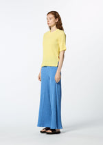 A person with long, wavy hair and a neutral expression stands against a white background, wearing HaaT's KYO CHIJIMI MAY textured yellow round-neck top in ultra-fine cotton, loose blue pants, and black sandals.