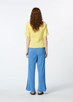 A person with long curly hair, seen from behind, wears the HaaT KYO CHIJIMI MAY yellow round-neck ultra-fine cotton top and loose blue pants with black flats, standing against a plain white background.