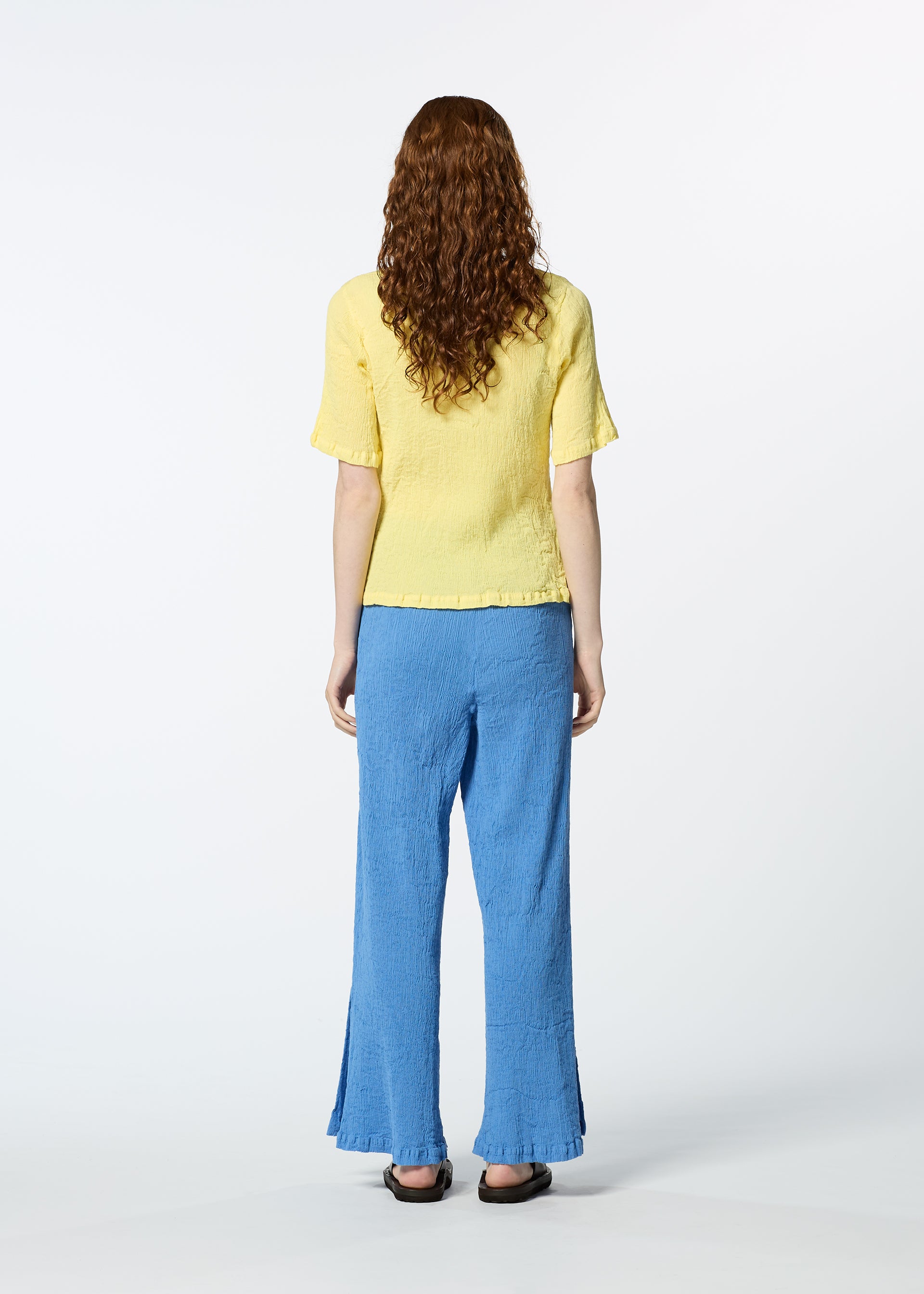 Zoomed view A person with long curly hair, seen from behind, wears the HaaT KYO CHIJIMI MAY yellow round-neck ultra-fine cotton top and loose blue pants with black flats, standing against a plain white background.