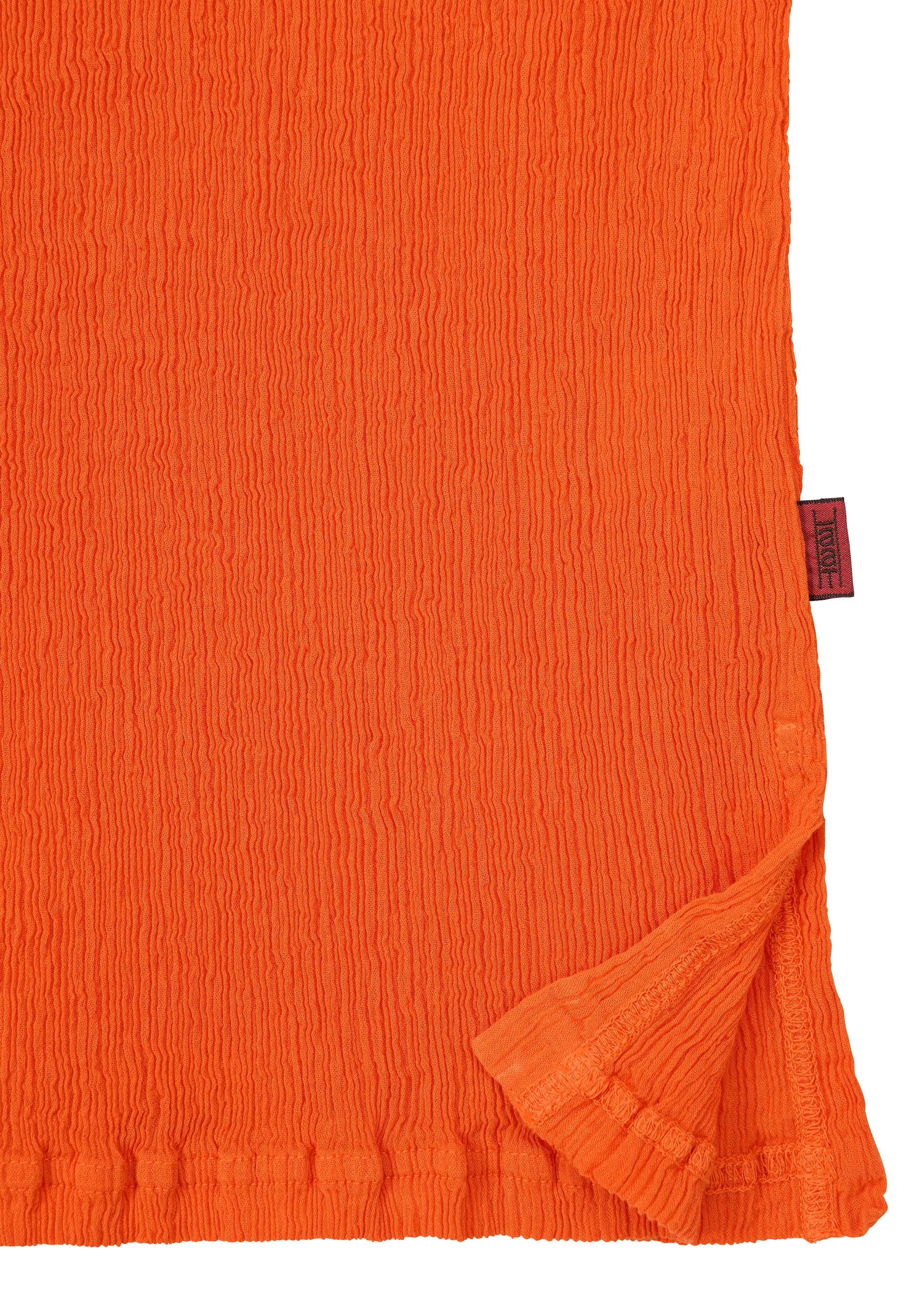 Zoomed view A close-up of the textured orange fabric from the HaaT KYO CHIJIMI MAY top, highlighting its ultra-fine cotton weave, slightly crinkled surface, a folded corner, and a small red tag on the edge.