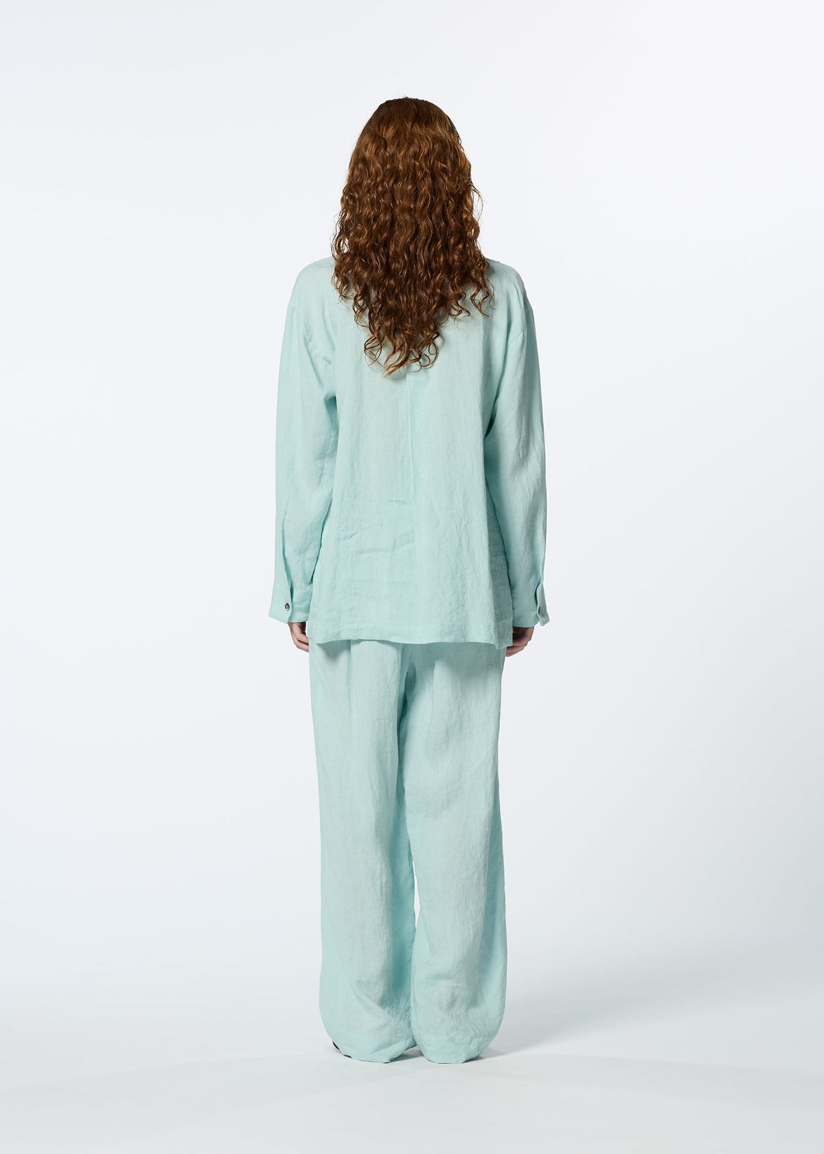 A person with long curly hair stands facing away from the camera, wearing HaaT's JIKI women's blouse and matching pants in light blue against a plain white background. view 8 of 8