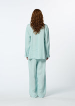 A person with long curly hair stands facing away from the camera, wearing HaaT's JIKI women's blouse and matching pants in light blue against a plain white background.