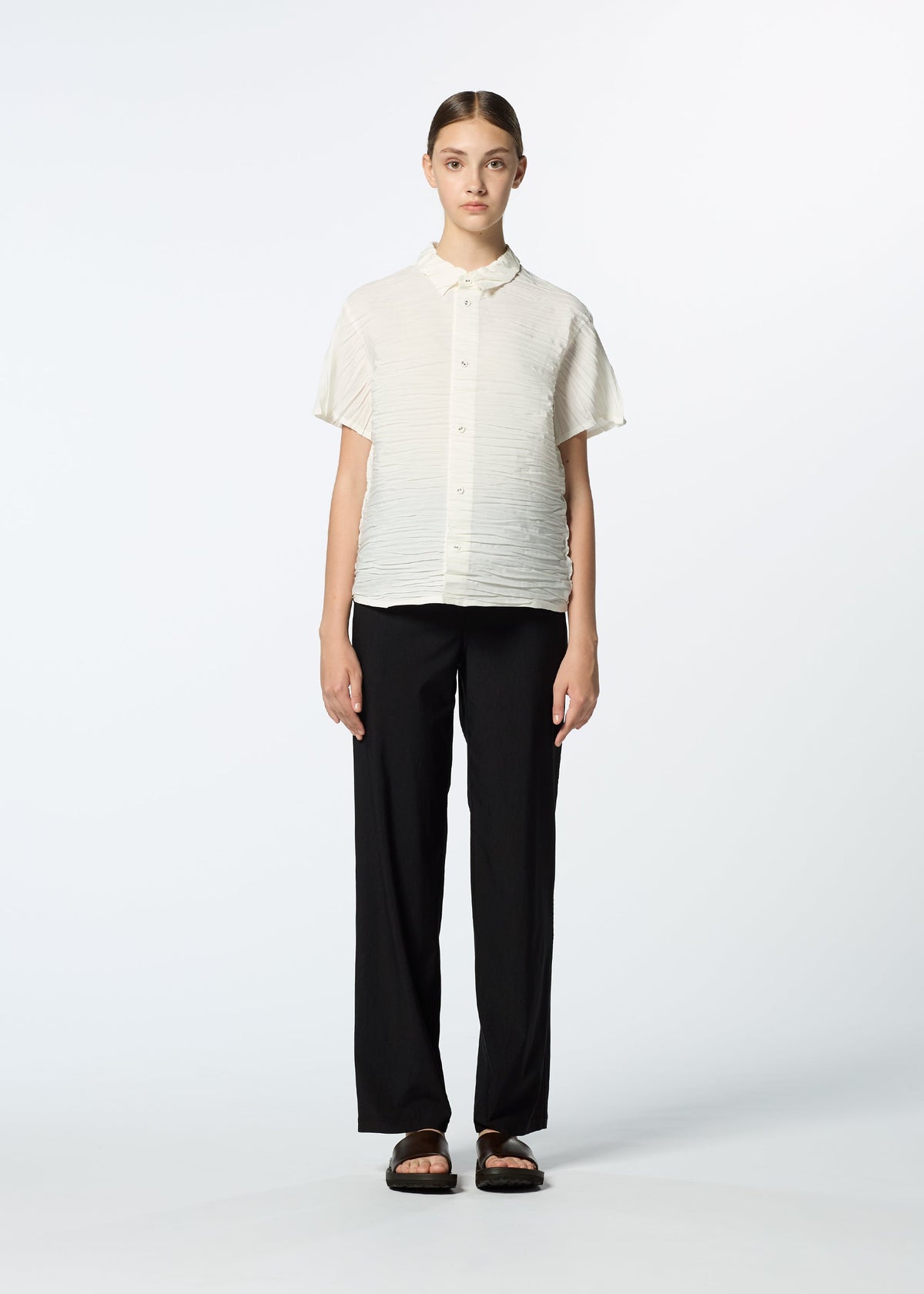 A person faces forward against a plain white background, wearing HaaT's SHIBARI SHIRT made from recycled polyester, loose black pants, and black sandals. Their hair is parted in the center and tied back. view 7 of 10