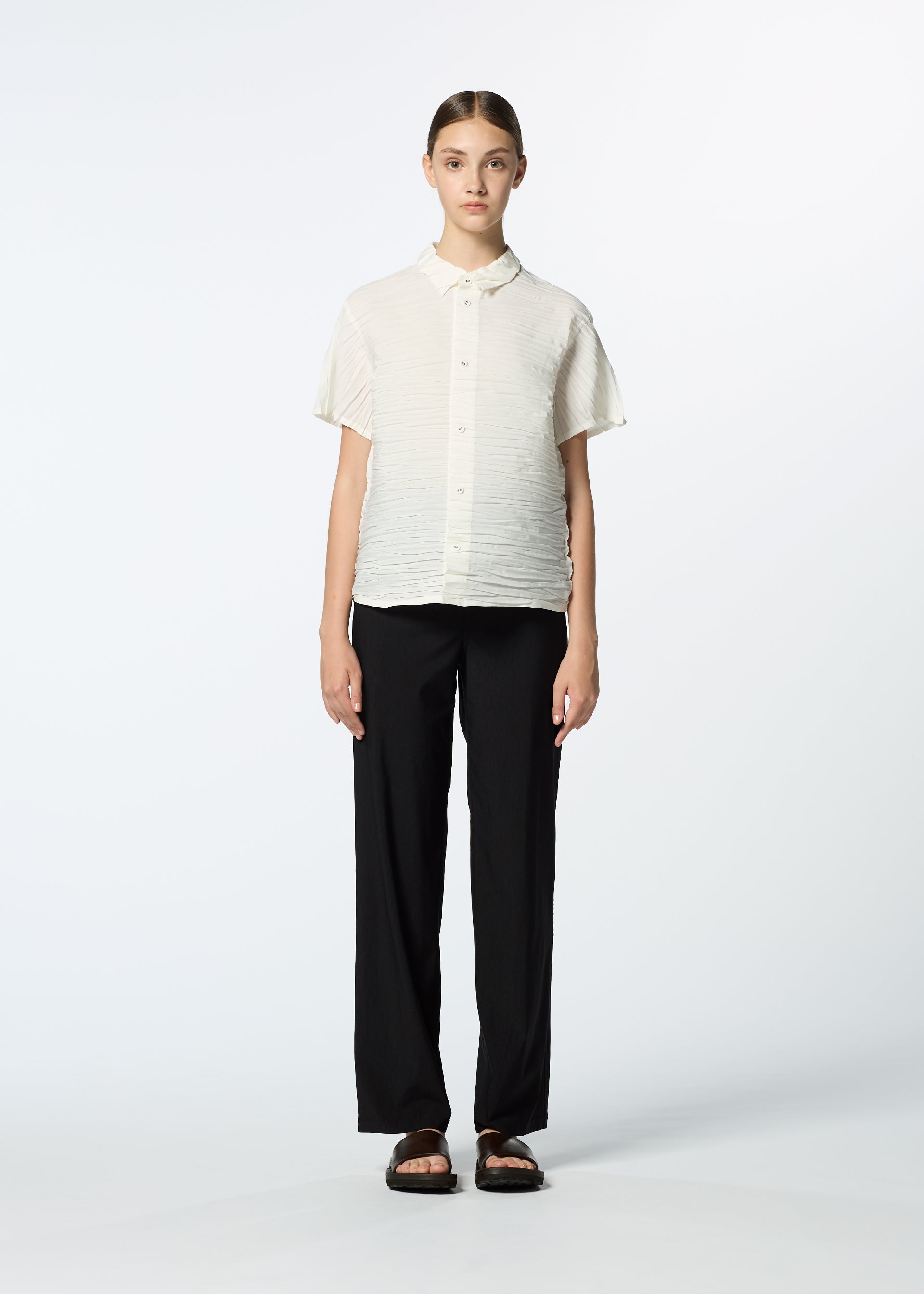 Zoomed view A person faces forward against a plain white background, wearing HaaT's SHIBARI SHIRT made from recycled polyester, loose black pants, and black sandals. Their hair is parted in the center and tied back.