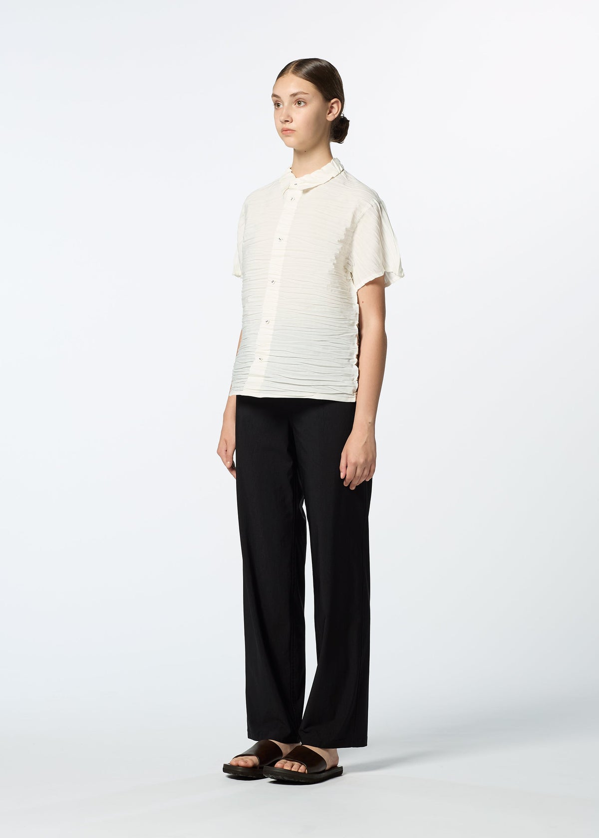 A woman stands against a white background wearing the HaaT SHIBARI SHIRT—a short-sleeved, textured white button-up made from recycled polyester—paired with loose black pants and black sandals. Her hair is pulled back as she looks forward neutrally. view 8 of 10