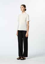A woman stands against a white background wearing the HaaT SHIBARI SHIRT—a short-sleeved, textured white button-up made from recycled polyester—paired with loose black pants and black sandals. Her hair is pulled back as she looks forward neutrally.