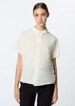 A young woman with straight brown hair pulled back wears the HaaT SHIBARI SHIRT, a short-sleeved top in recycled polyester, paired with black pants, standing against a plain light background.