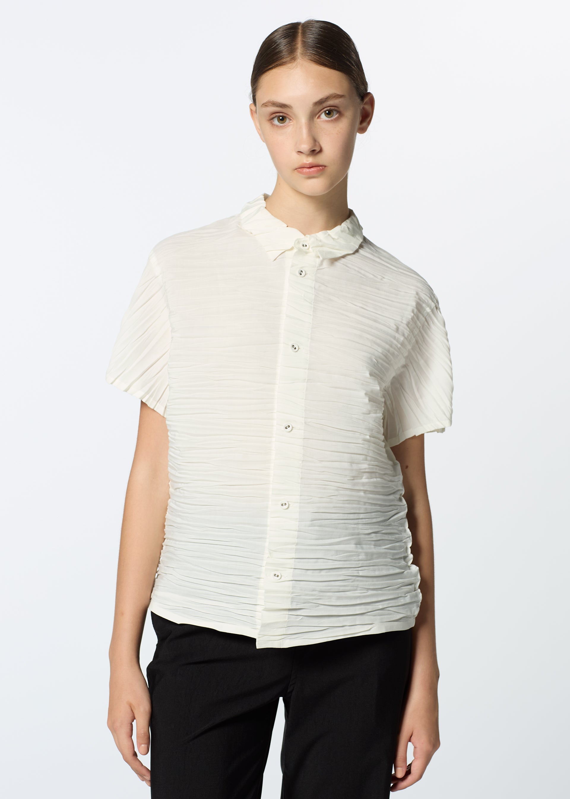 Zoomed view A young woman with straight brown hair pulled back wears the HaaT SHIBARI SHIRT, a short-sleeved top in recycled polyester, paired with black pants, standing against a plain light background.