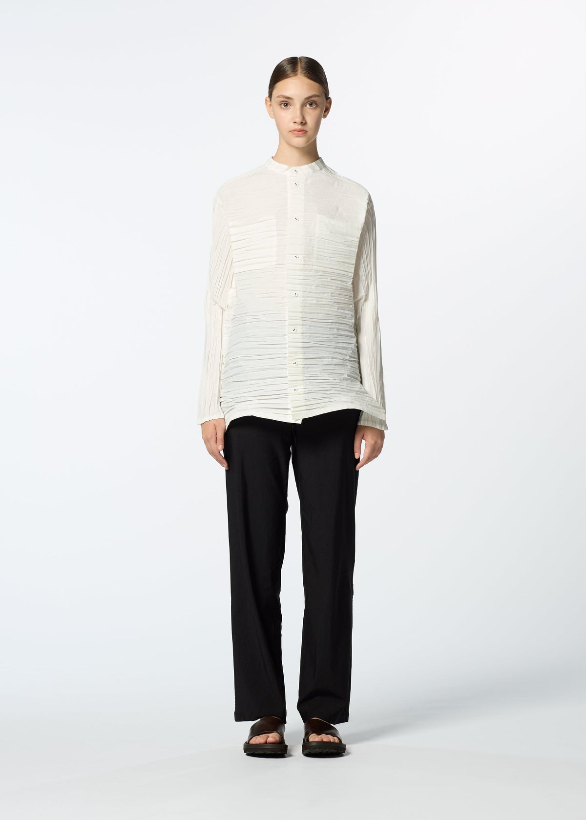 A person stands facing forward against a plain, light background, wearing HaaT’s SHIBARI SHIRT—a long-sleeved white button-up with subtle Shibari finish—paired with loose black pants and black sandals. Their hair is neatly parted and tied back. view 7 of 10