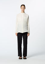 A person stands facing forward against a plain, light background, wearing HaaT’s SHIBARI SHIRT—a long-sleeved white button-up with subtle Shibari finish—paired with loose black pants and black sandals. Their hair is neatly parted and tied back.