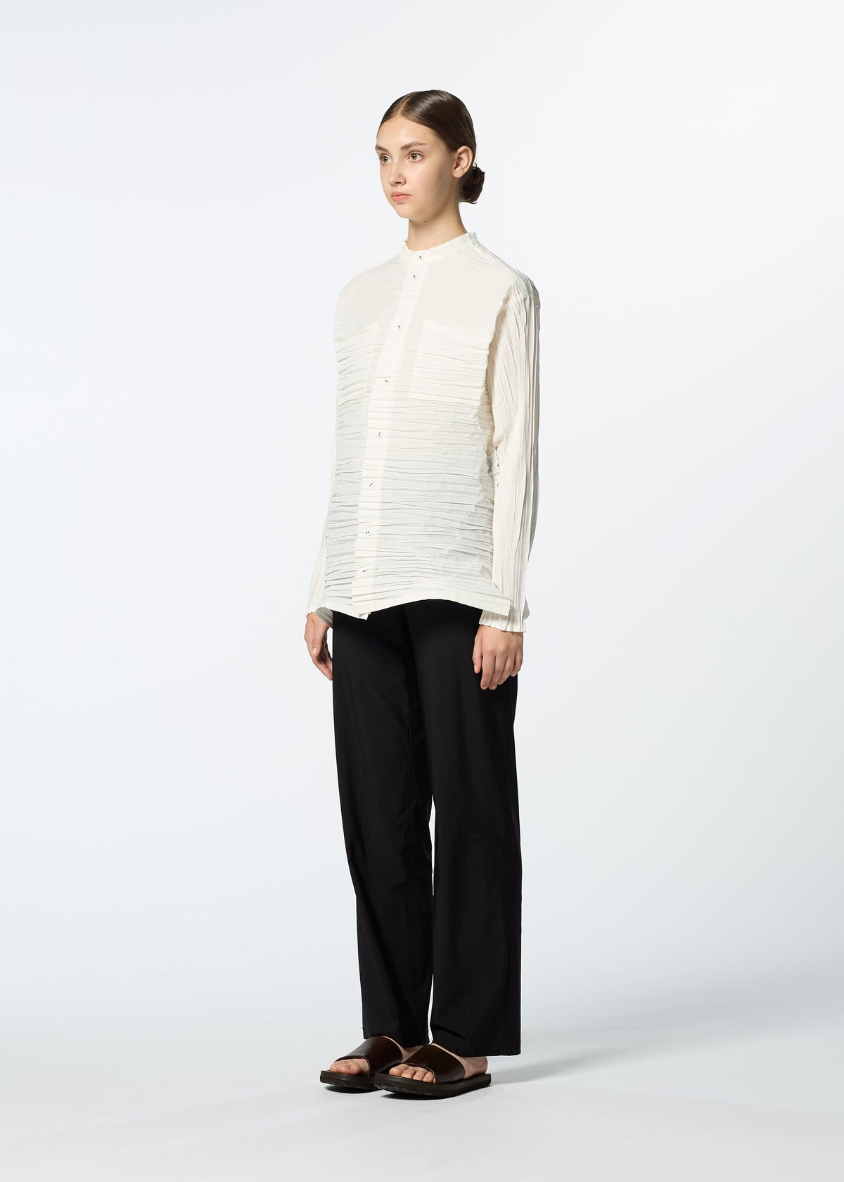 A person stands against a plain white background, wearing HaaT's SHIBARI SHIRT with subtle horizontal pleats, loose black pants, and black sandals. Their hair is tied back, and they gaze slightly to the side. view 8 of 10