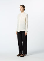 A person stands against a plain white background, wearing HaaT's SHIBARI SHIRT with subtle horizontal pleats, loose black pants, and black sandals. Their hair is tied back, and they gaze slightly to the side.