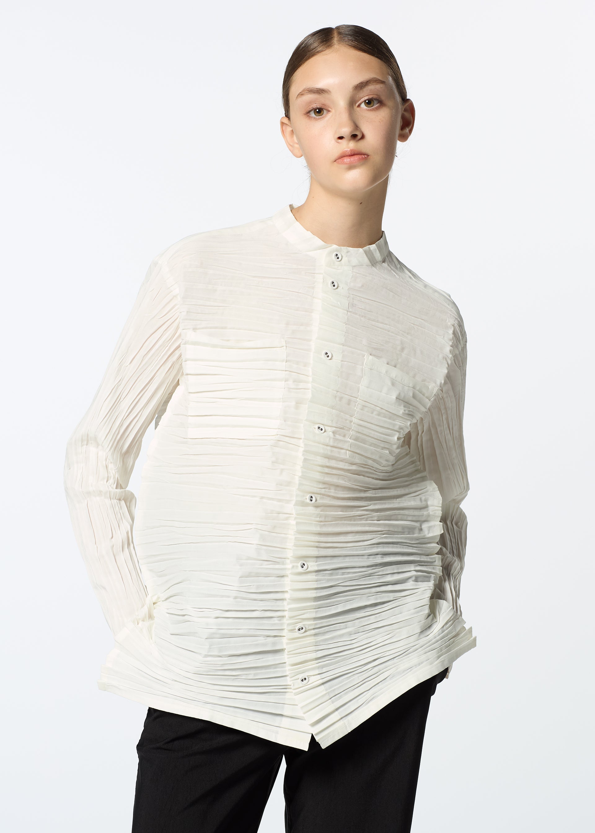 Zoomed view A young woman with brown hair slicked back wears the HaaT SHIBARI SHIRT, a white, long-sleeved top made from recycled polyester, paired with black pants. She stands against a plain white background with a neutral expression.