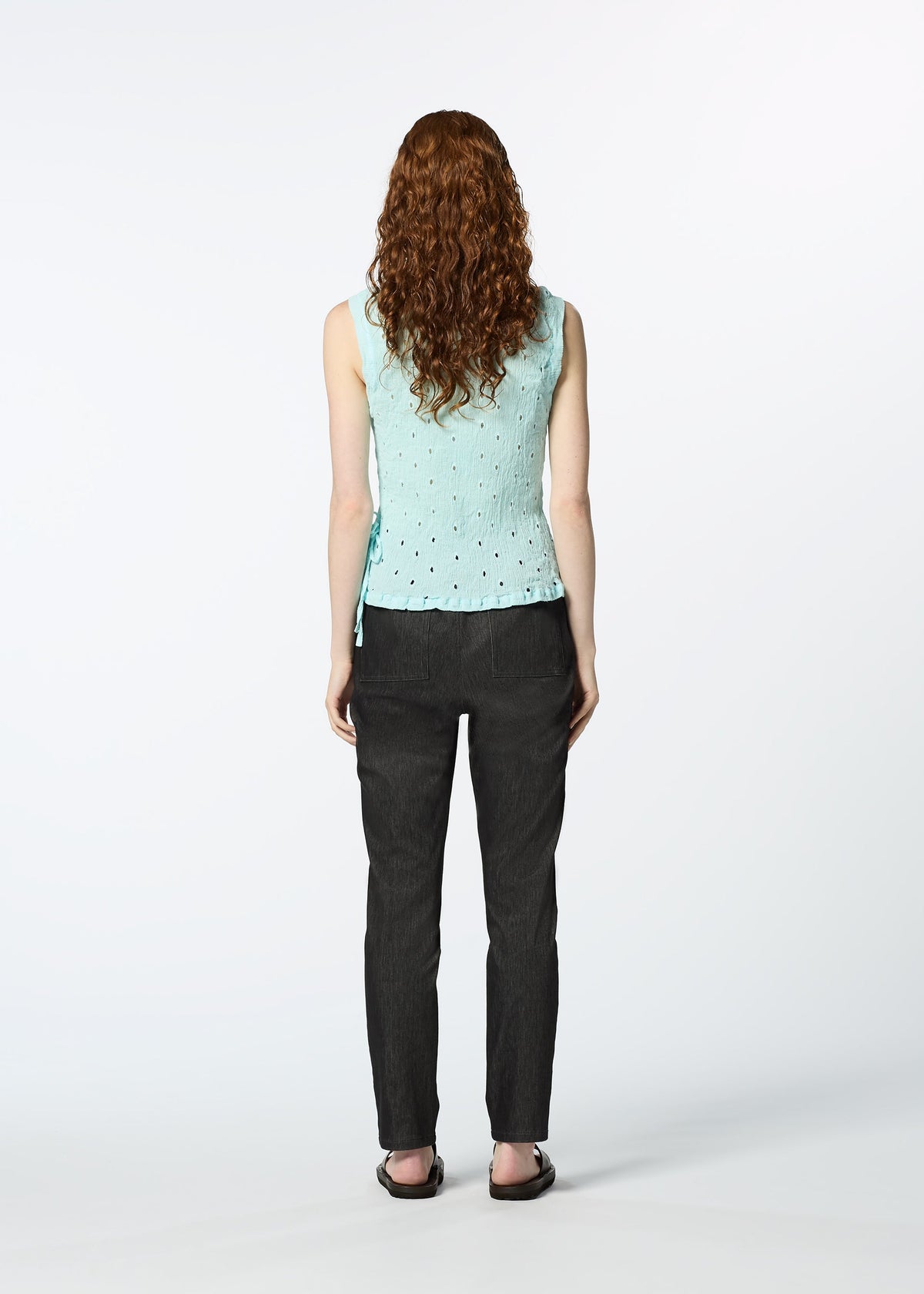 A person with long curly hair, dressed in a light blue HaaT KYO CHIJIMI TOBIKANNA CADEAU sleeveless top, dark pants, and black sandals, stands facing away against a plain white background. view 10 of 10