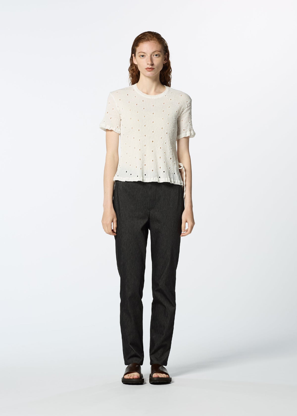 A person with long, wavy hair faces forward, wearing a white HaaT KYO CHIJIMI TOBIKANNA CADEAU short-sleeve top, dark straight-leg pants, and black sandals against a plain light background. view 8 of 11