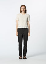 A person with long, wavy hair faces forward, wearing a white HaaT KYO CHIJIMI TOBIKANNA CADEAU short-sleeve top, dark straight-leg pants, and black sandals against a plain light background.