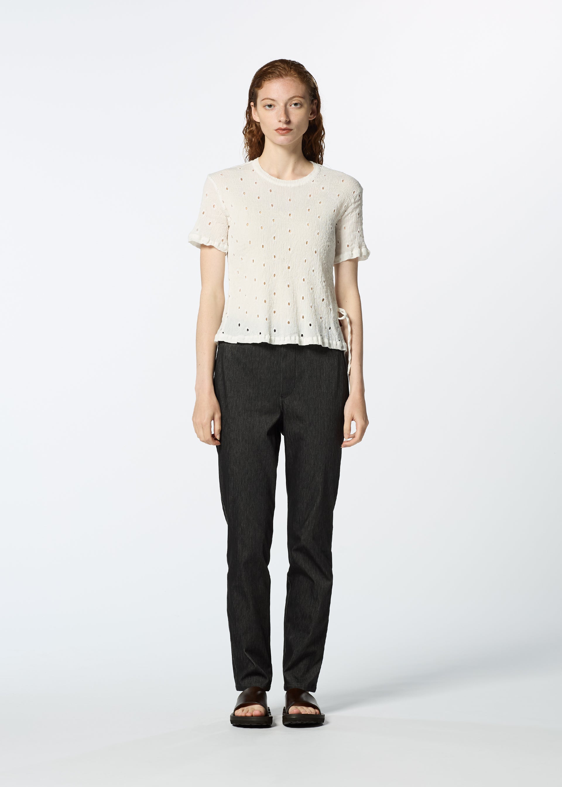 Zoomed view A person with long, wavy hair faces forward, wearing a white HaaT KYO CHIJIMI TOBIKANNA CADEAU short-sleeve top, dark straight-leg pants, and black sandals against a plain light background.