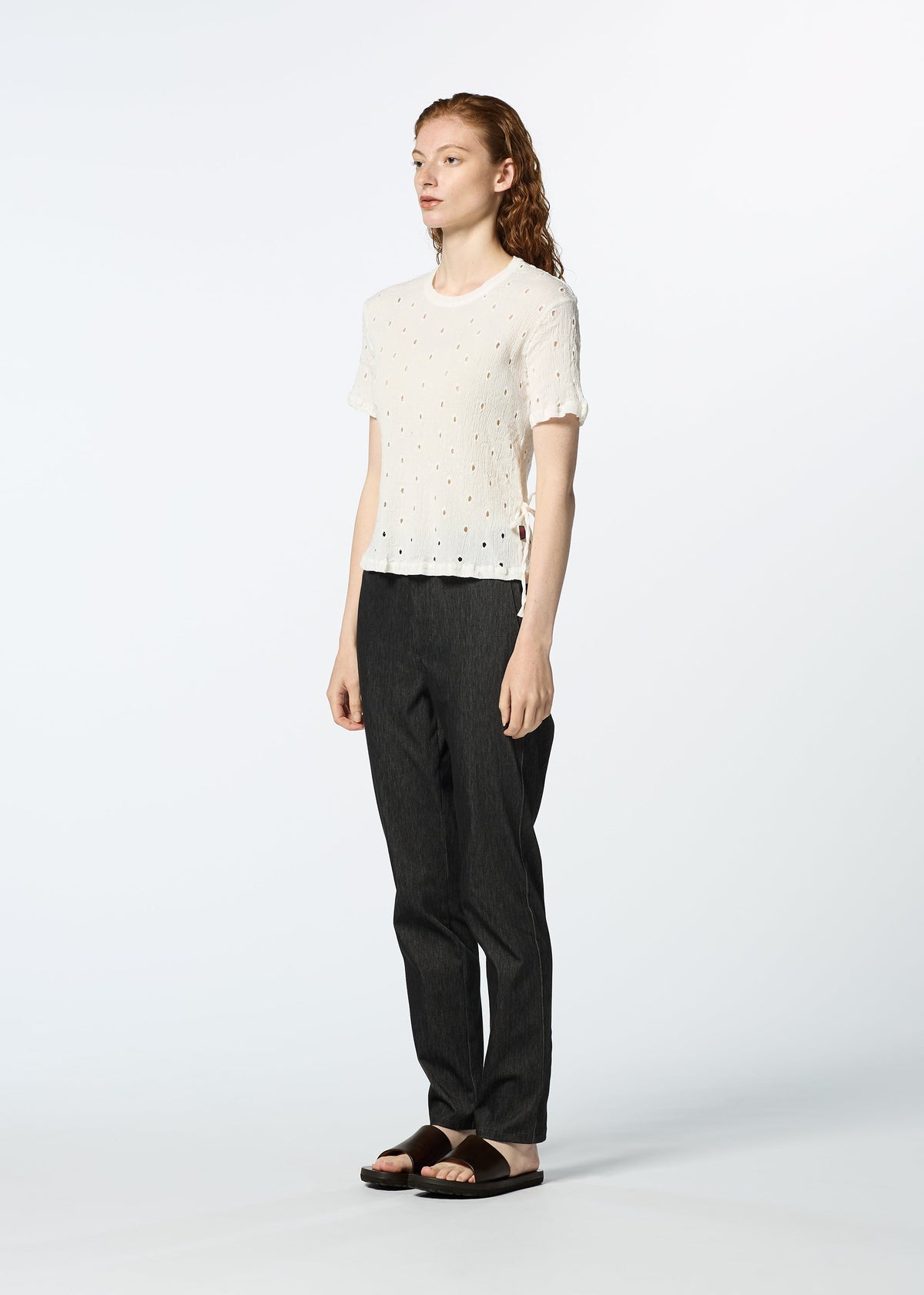 A person with long hair, facing slightly to the side, wears a white HaaT KYO CHIJIMI TOBIKANNA CADEAU short-sleeve top, dark pants, and black sandals against a plain light background. view 9 of 11