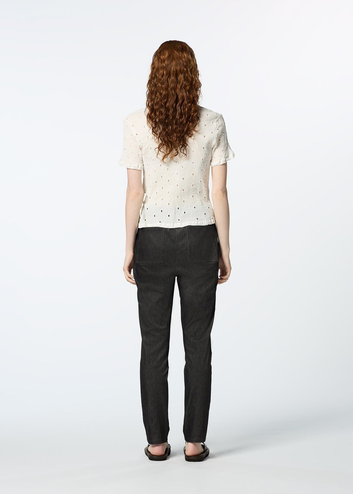 A person with long, curly hair stands facing away, wearing the HaaT KYO CHIJIMI TOBIKANNA CADEAU short-sleeve top, dark pants, and flat shoes against a plain white background. view 10 of 11