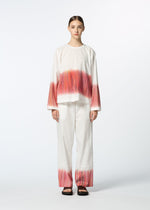 A person stands facing forward in HaaT’s SAZANAMI—an organic cotton ensemble featuring a loose fit and hand-painted red and orange brushstroke patterns. They wear black sandals, have straight hair, and a neutral expression.