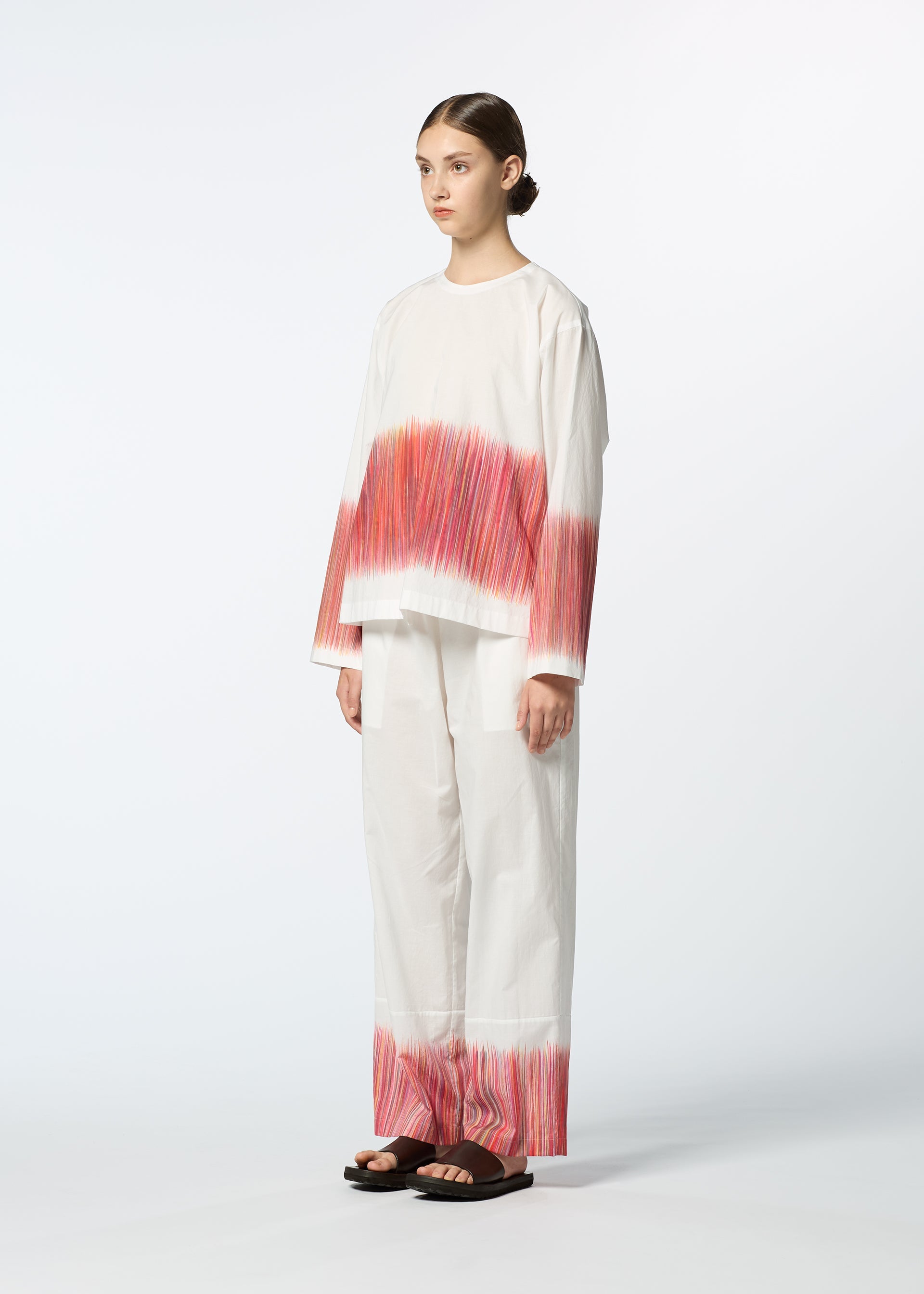 Zoomed view A person stands against a plain white background wearing HaaT's SAZANAMI, a loose organic cotton outfit with red and pink hand-painted horizontal streaks by Kyoto artisans on the sleeves, lower top, and pant legs, paired with black sandals.