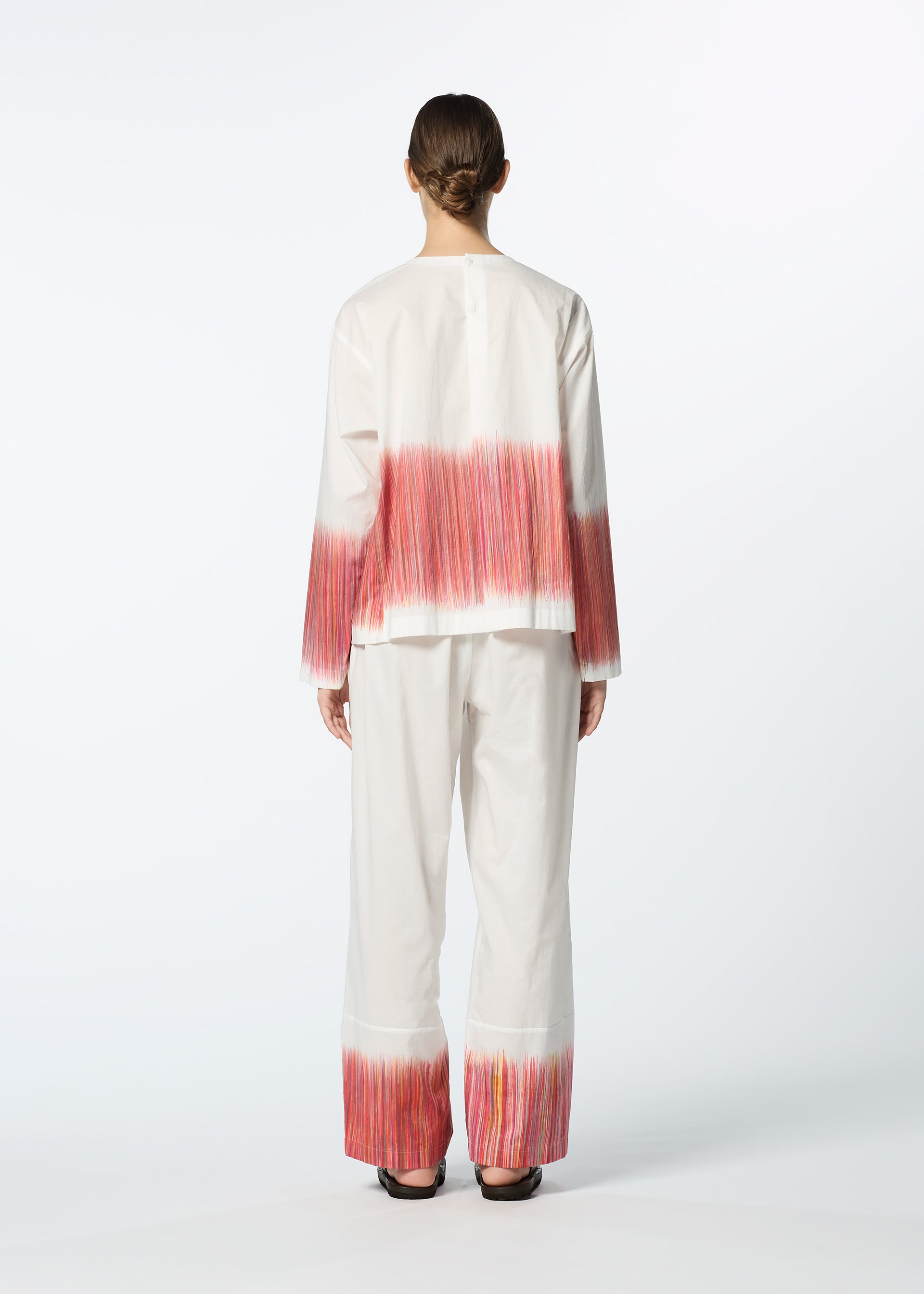 Zoomed view A person faces away, wearing the HaaT SAZANAMI top—an organic cotton piece hand-painted with vibrant pink and orange horizontal brushstrokes on the back and cuffs. The background is plain white.